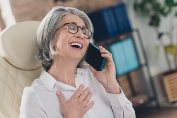 Portrait of excited overjoyed aged person sit chair chatting telephone hand chest laughing workplace indoors