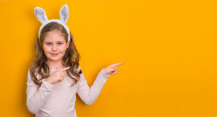 Little girl with easter bunny ears pointing on empty yellow background with copy-space.