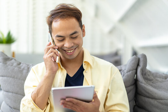 smiling Asian young businessman answering over cellphone and searching for information on the internet with digital tablet, enjoy chatting online in social media mobile applications with friends