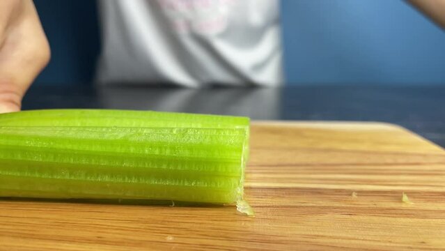 Female hands using knife to cut raw organic celery slice on a wooden chopping board at home kitchen close up shot. Healthy lifestyle, fresh vegetable and cooking concept