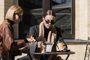stylish young couple eating asian food from carton boxes near coffee to go on bistro table.