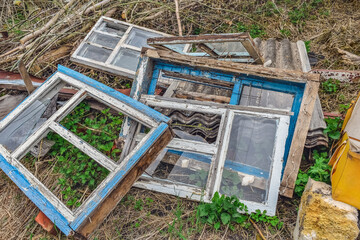 A pile of old wooden windows with broken glass lies on the ground with young nettles and grass in spring. Dismantling for building materials of old houses in Ukraine