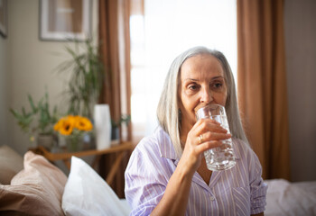 Portrait of senior woman sitting on the bed in pajamas and drinking water.