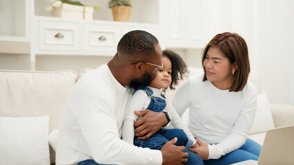 Multiracial newbie parent is sitting on a sofa with their daughter and giving love to her while watch online entertainment contents on a laptop. African adult man is kissing his daughter to show love.