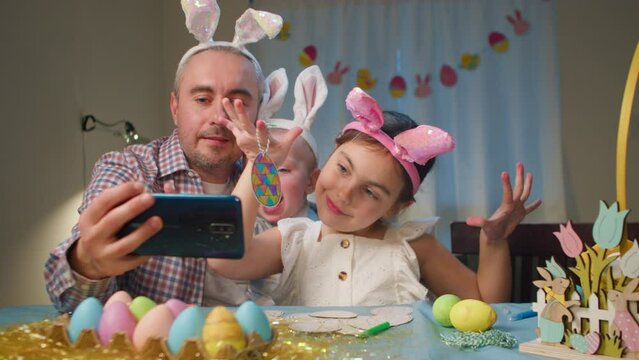 Happy family with bunny ears on head at home at table smiling and posing at smartphone camera while dad takes selfie photo. Father, Daughter and little son are photographed together celebration Easter