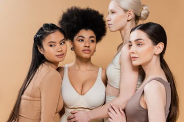 Obraz premium sensual multicultural women in underwear touching each other while posing isolated on beige.