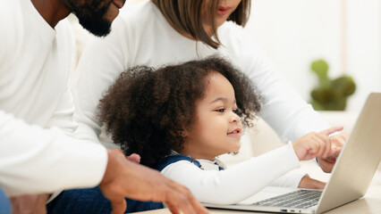 Obraz premium Close-up of a curious daughter touching a laptop screen to select online kid contents while multiracial newbie parent taking care of her. A happy family is teaching their daughter to use a laptop.