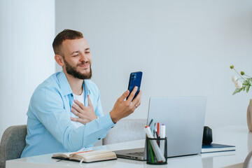 Handsome beardy caucasian young man sitting at table with laptop makes video call by phone touches...