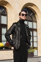 Fototapeta na wymiar tattooed man in stylish sunglasses and black leather jacket standing with hands in pockets on urban street.