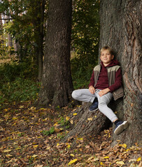 A handsome guy sits on the root of a huge oak tree leaning against the trunk of a tree and smiles.