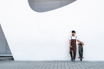 Young man wearing headphones standing by skateboard in front of wall