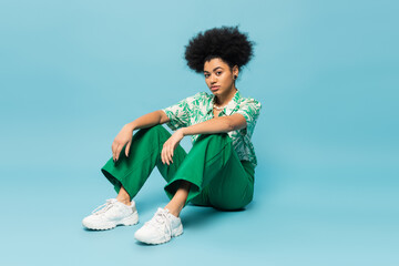 full length of curly african american woman in green pants and white sneakers sitting on blue background.