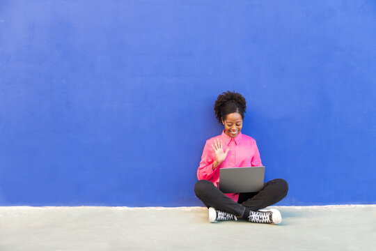 Woman waving on video call through laptop sitting in front of blue wall