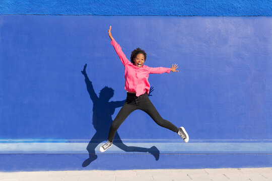 Carefree woman jumping on footpath in in front of blue wall