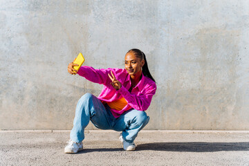 Smiling young dancer in squatting position taking selfie through mobile phone in front of wall