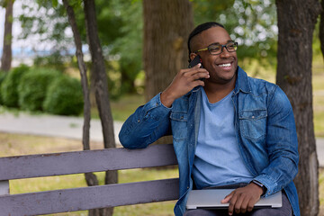Online learning in a convenient location for an international student with a laptop in hand. Smiling black man shares his academic progress over the phone to relatives sitting on a bench in the park