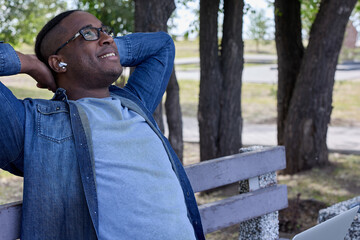 Fototapeta premium On his summer vacation black office worker relaxes in park enjoying nature and silence. An African student in glasses and headphones relaxes listening to his favorite audio channel sitting on a bench