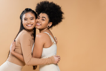 joyful asian and african american women in underwear embracing and looking at camera isolated on beige.