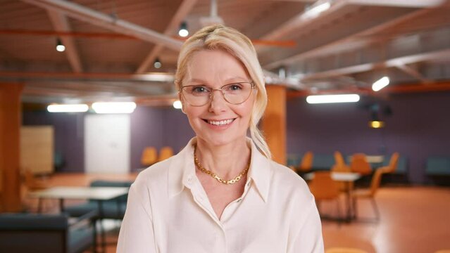 Close-up portrait of nice-looking mature beautiful businessperson in stylish eyewear. Sliming blonde woman 50s looking into camera at modern office background. Career people. Elegant lady 50s portrait