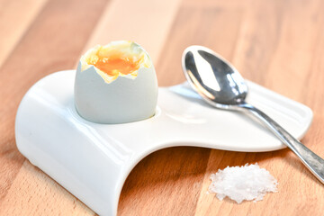a single boiled egg in an egg cup broken open showing yolk with a spoon on a wooden surface - 576032332