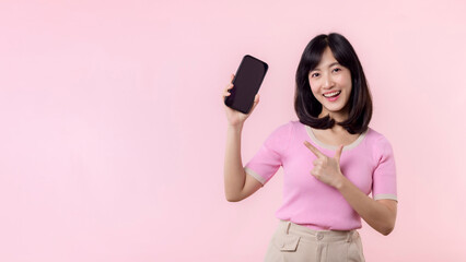 Portrait beautiful young asian woman happy smile showing smartphone screen with pointing finger hand gesture to mobile phone isolated on pink pastel studio background. Blank screen smartphone concept