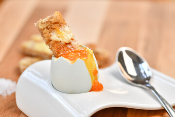 a single boiled egg in an egg cup, broken open showing yolk and with a slice of toast inserted with a spoon on a wooden surface - 576032999