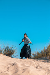 A Japanese warrior in a traditional kimano armed with a katana sword on a sandy shore next to the ocean during sunset. A man from medieval Asia. Reconstruction of cultural heritage. Culture in Japan. 