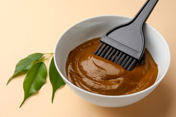 Bowl of henna cream, brush and green leaves on beige background, closeup. Natural hair coloring