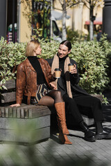 full length of happy couple holding paper cups with coffee to go while sitting on bench.