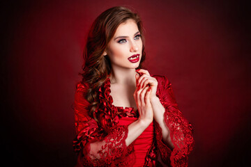 Portrait of a young, attractive vampire woman in a red rococo dress posing isolated against a dark background with red backlights.