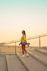 Pretty young woman with brown skin with long black braids standing holding a skateboard with her hand dressed in warm colored summer clothes on a concrete stairs on the seafront at summer sunset.