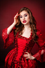 Portrait of a young, attractive vampire woman in a red rococo dress posing isolated against a dark background with red backlights.