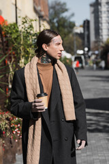 young tattooed man in coat and scarf holding paper cup with coffee to go.