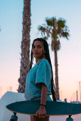Pretty young woman with brown skin with long black braids posing with her skateboard dressed in a...