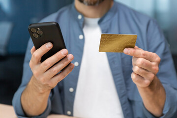 Close-up of man's hands inside the office, businessman in blue shirt and white t-shirt works at workplace, man holds bank credit card and smartphone, makes online purchase in online store.