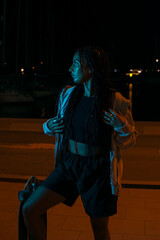 Naklejka premium Pretty young woman with brown skin with long black braids holding her skateboard dressed in a gray sweatshirt and dark clothes posing on the seafront on a summer night with blue led light.