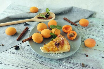 A piece of apricot pie on a plate, sprinkled with powdered sugar, spices and mint leaves, fresh apricots on the kitchen table, homemade food, sweet dessert