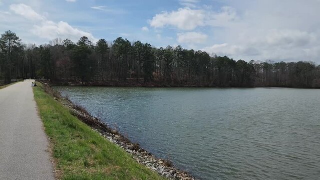 footage along the banks of Lawrenceville Lake with people sitting on the grass and a woman walking along the footpath wearing a pink shirt at Rhodes Jordan Park in Lawrenceville Georgia USA