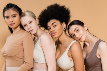 young multiethnic women in underwear leaning on each other and looking at camera isolated on beige.