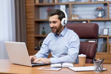 Mature businessman with beard and shirt working contentedly in office sitting at desk, man in headphones listening to podcasts and audiobooks at workplace, boss typing on laptop.