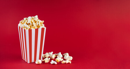 Red white striped carton bucket with tasty cheese popcorn, isolated on red background. Box with scattering of popcorn grains. Fast food, movies, cinema and entertainment concept.