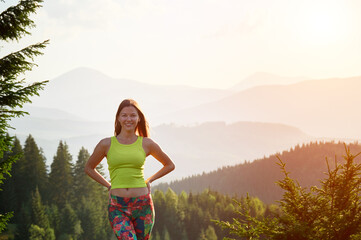 Portrait of young attractive woman posing outdoors at sunset. Slim, sporty female tourist smiling, admiring landscape, wearing sportswear. Concept of traveling and hiking in mountains.