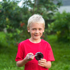 blond boy is smiling in a red T-shirt, holding a caliper and measuring something. natural light. background foliage of trees