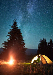 Night camping in mountains under starry sky and Milky way. Female tourist sitting in tent entrance...