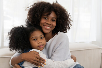 Happy afro family embracing together on sofa at home. Smiling African American young mother hugging her child daughter while sitting on couch at home