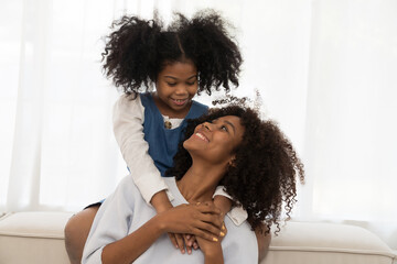 Happy afro family embracing together on sofa at home. Smiling African American young mother hugging...