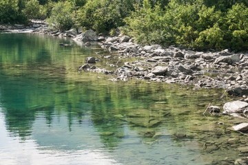 Reflection of treetops and mountain ranges in a turquoise-colored mountain lake with a rocky shore, a landscape on a sunny summer day in the mountains