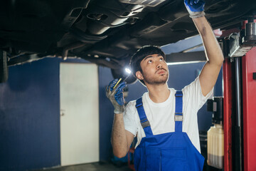 Asian mechanic or repairman using flashlight checking under car broken part at auto repair shop....