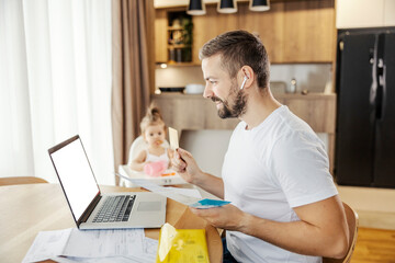 A happy father is paying bills online on a laptop while babysitting his daughter.