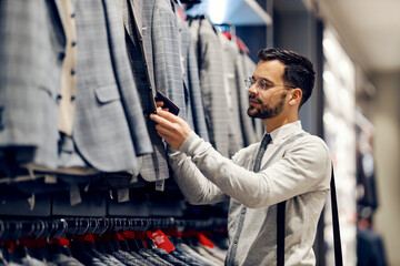 A young elegant man is standing at clothing store and looking for proper suit.